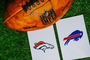 An NFL football sits on a grass field with two white cards depicting the Denver Broncos' and Buffalo Bills' logos.